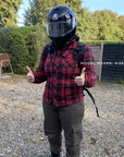 Person wearing a red plaid shirt, black helmet, and backpack giving a thumbs-up gesture outdoors.
