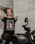 Woman sitting on a Motorcycle wearing a T-shirt with a women wearing a Motorcyclehelmet print and with the text "Wild dust sisters woman rider" and "Speed Goddess"