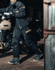 Person in dark clothing with gold accents standing next to a motorcycle in a rustic setting