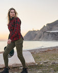 Woman in plaid shirt and green pants standing by a body of water with mountains in the background.