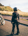 Person in motorcycle gear standing next to a motorcycle on a road with a scenic background