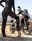 A woman standing by her motorcycle wearing Black SHIMA touring motorcycle pants for women