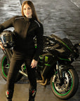 A young woman standing by the motorcycle wearing black motorcycle protective clothes