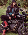 A woman leaning on her Triumph motorcycle wearing black leather motorcycle jacket and red boots