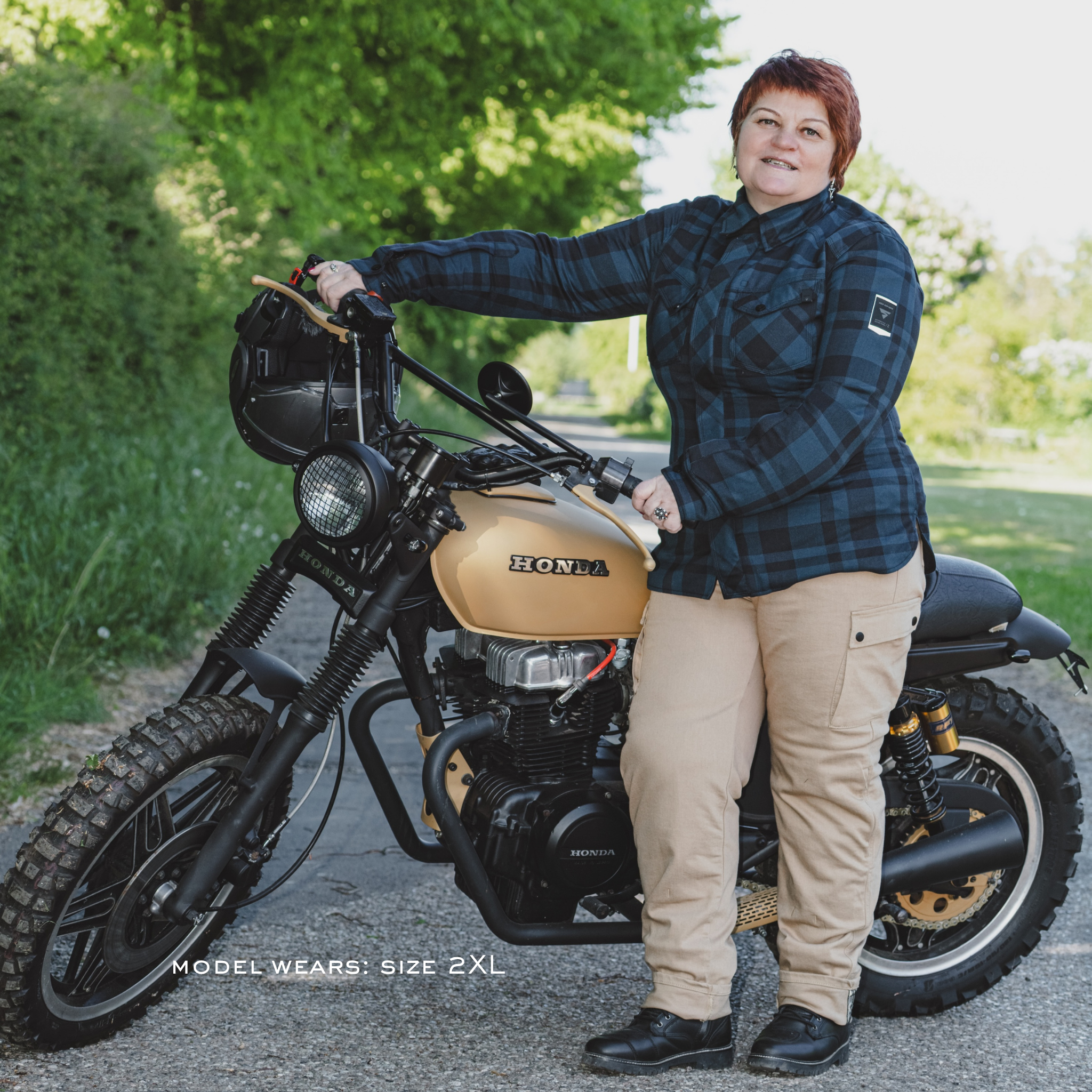 a woman standing next to a Honda motorcycle on a road with greenery in the background wearing blue lumberjack motorcycle shirt from Shima