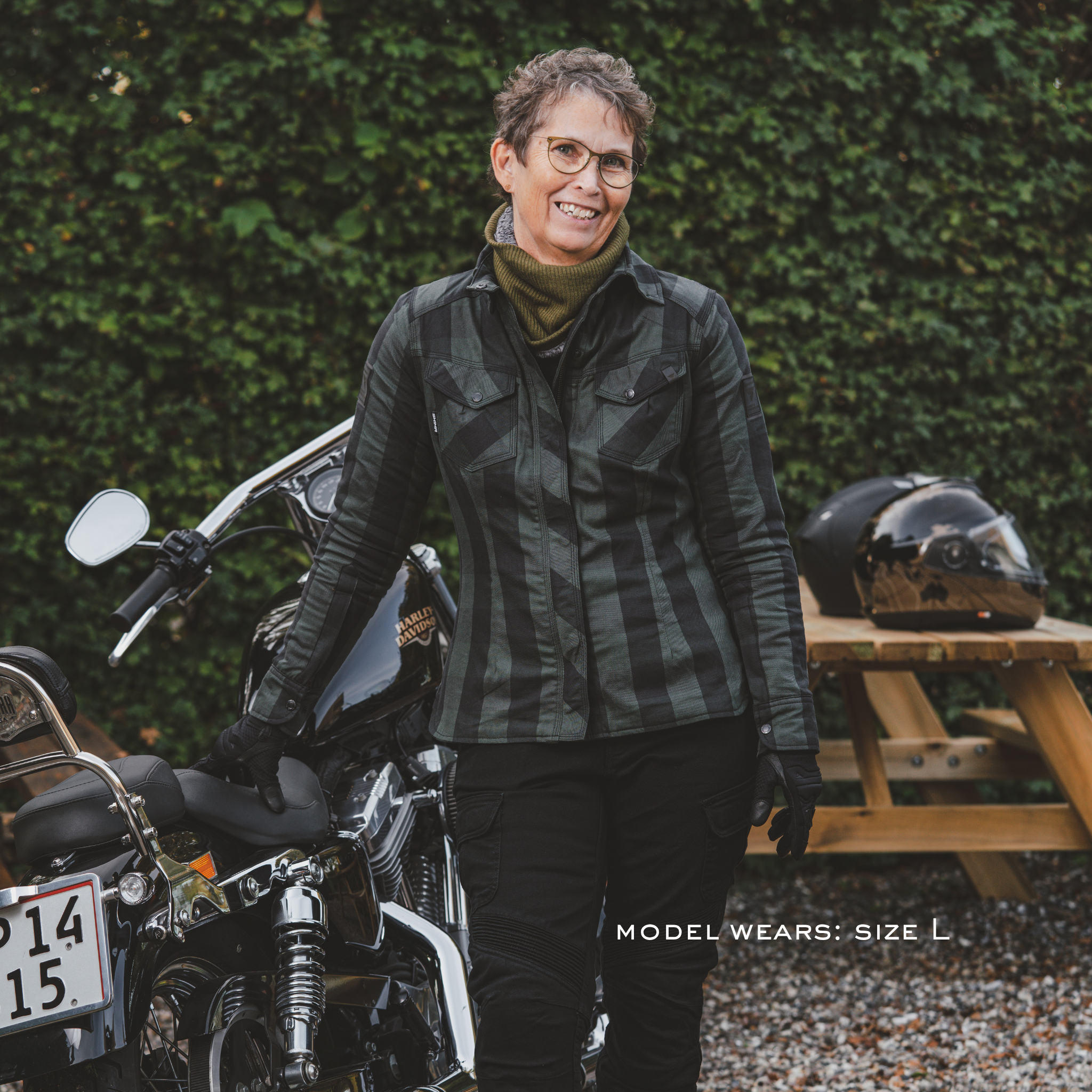 Person wearing a plaid jacket standing next to a motorcycle with a helmet on a table in the background.