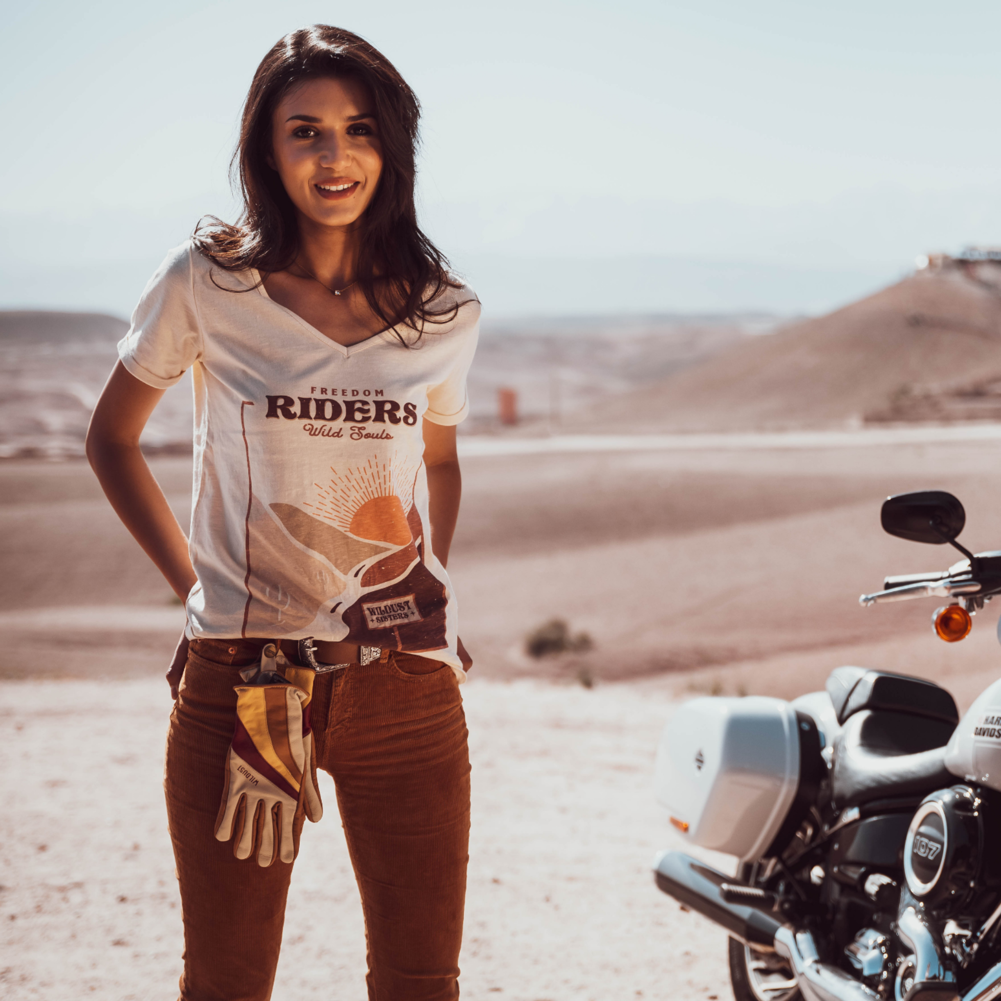 Woman standing in the dessert next to a Motorcycle wearing a T-shirt with a sunset/sunrise print and the text "Freedom Riders Wild Souls" and "Wildust sisters"