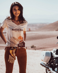 Woman standing in the dessert next to a Motorcycle wearing a T-shirt with a sunset/sunrise print and the text "Freedom Riders Wild Souls" and "Wildust sisters"