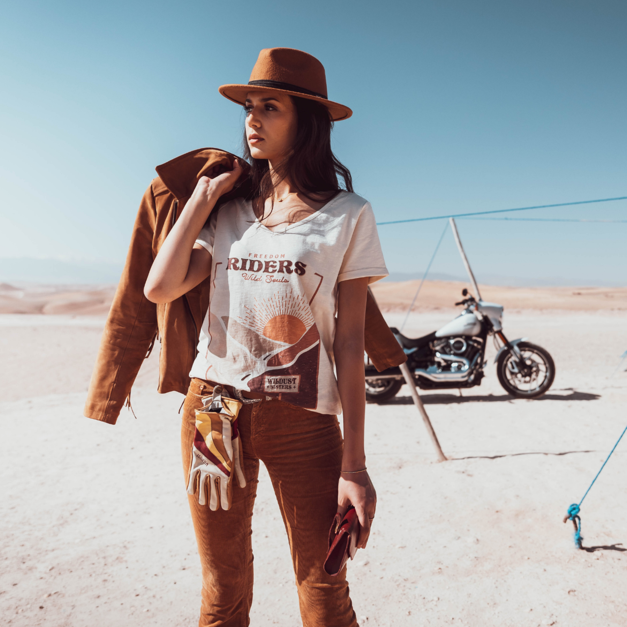 Woman standing in the dessert wearing a T-shirt with a sunset/sunrise print and the text "Freedom Riders Wild Souls" and "Wildust sisters"