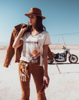 Woman standing in the dessert wearing a T-shirt with a sunset/sunrise print and the text "Freedom Riders Wild Souls" and "Wildust sisters"