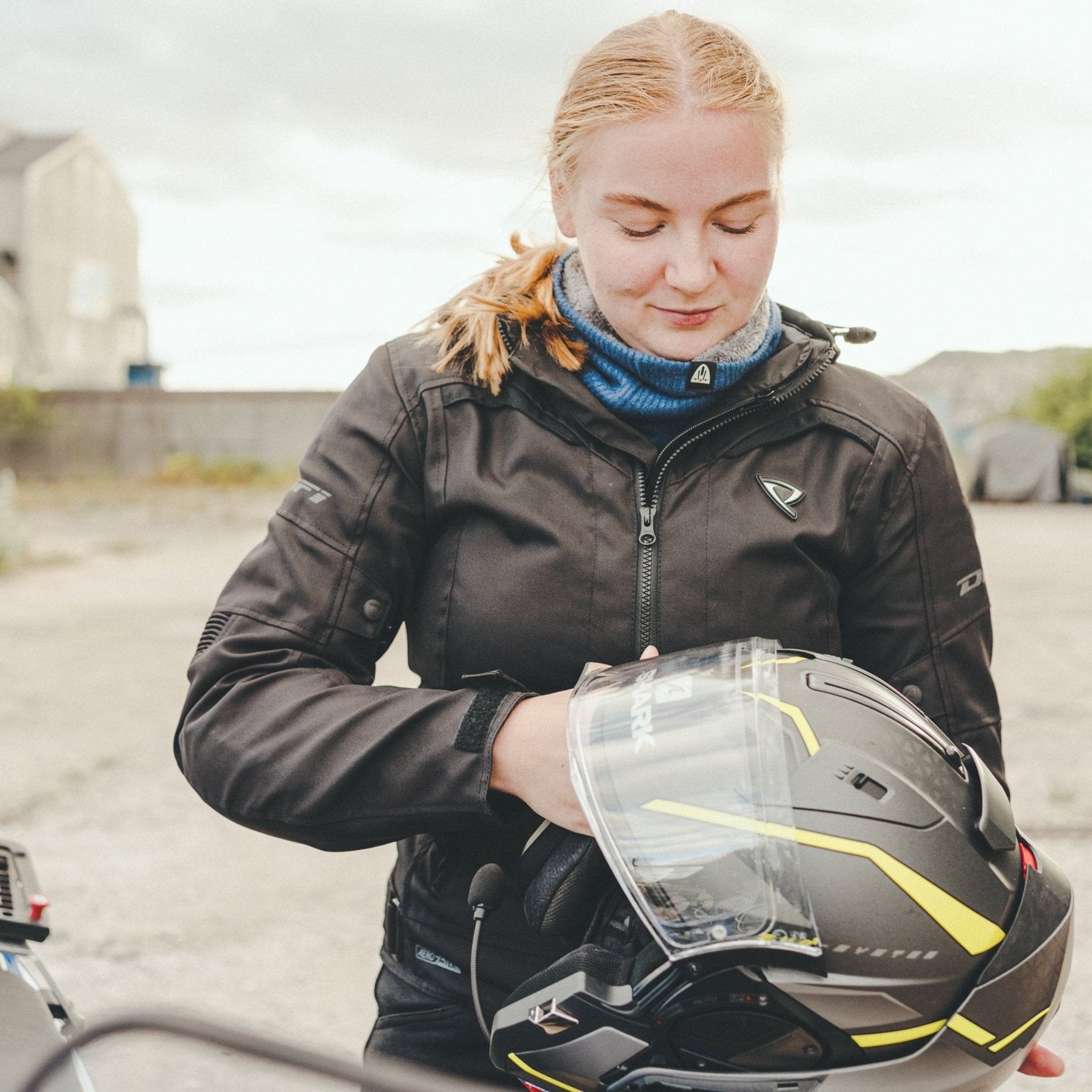 light red-haired woman wearing a blue knitted neck warmer from Moto Girl, standing with a helmet in her hands.