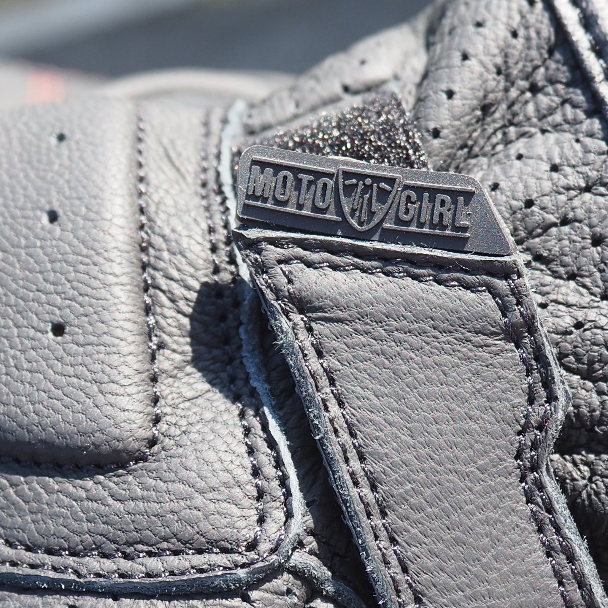 Close-up of a gray leather glove with a MotoGirl brand logo on a blurred background