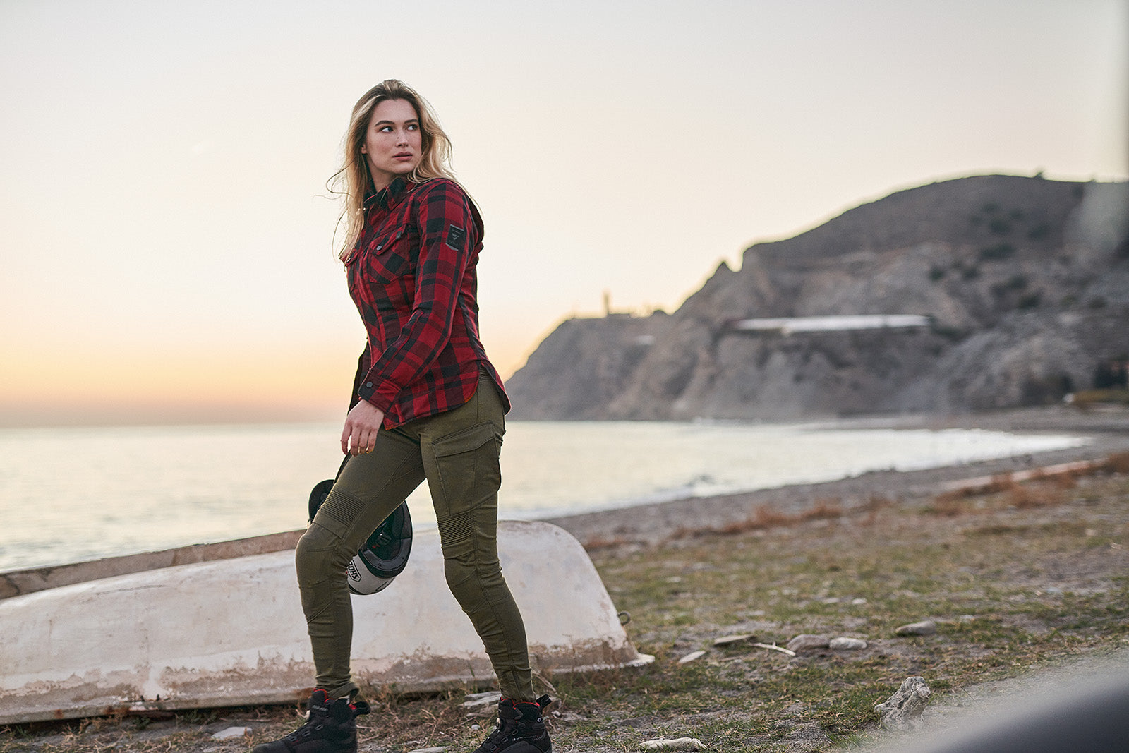Woman in plaid shirt and green pants standing by a body of water with mountains in the background.