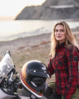 Woman in a plaid RED motorcycle shima jacket standing next to a motorcycle with a helmet on the seat, outdoors.