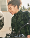 Woman sitting on a motorcycle, wearing a green plaid shirt, with a blurred background.