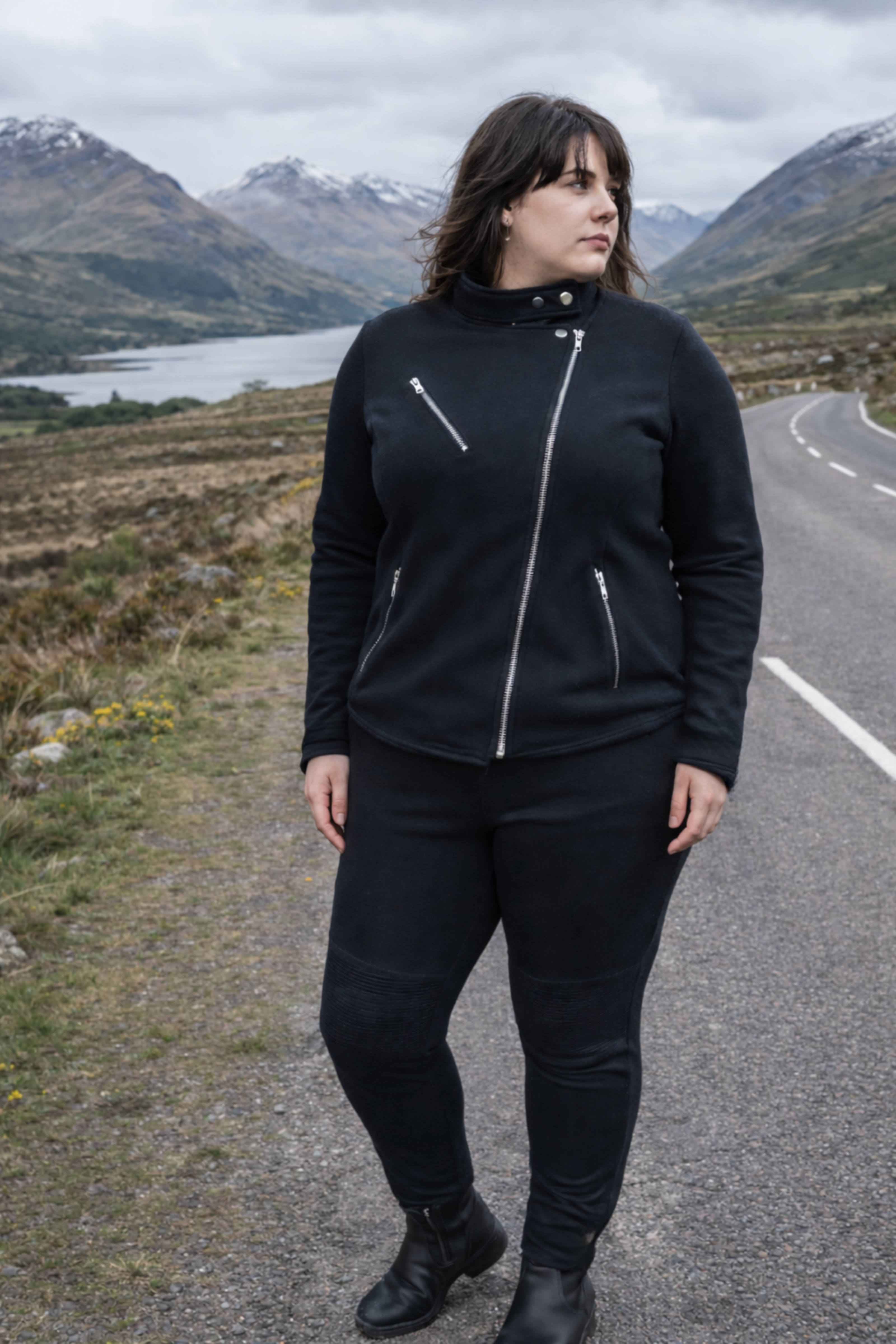 Person wearing a black outfit standing on a road with mountains and a lake in the background
