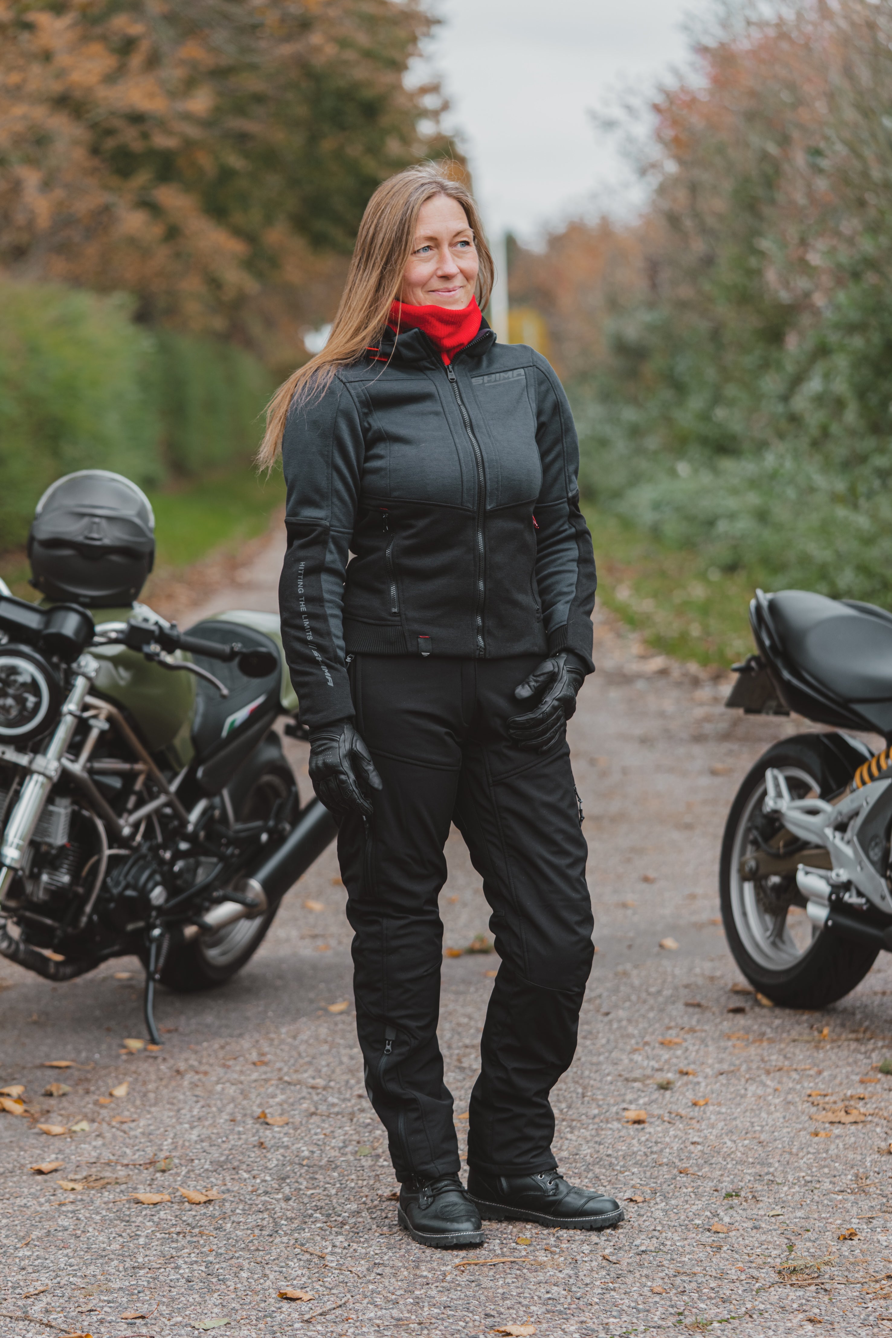 A woman wearing a black motorcycle jacket, black pants, and a bright red neck gaiter stands on a gravel path between two motorcycles in an outdoor setting with autumn trees.