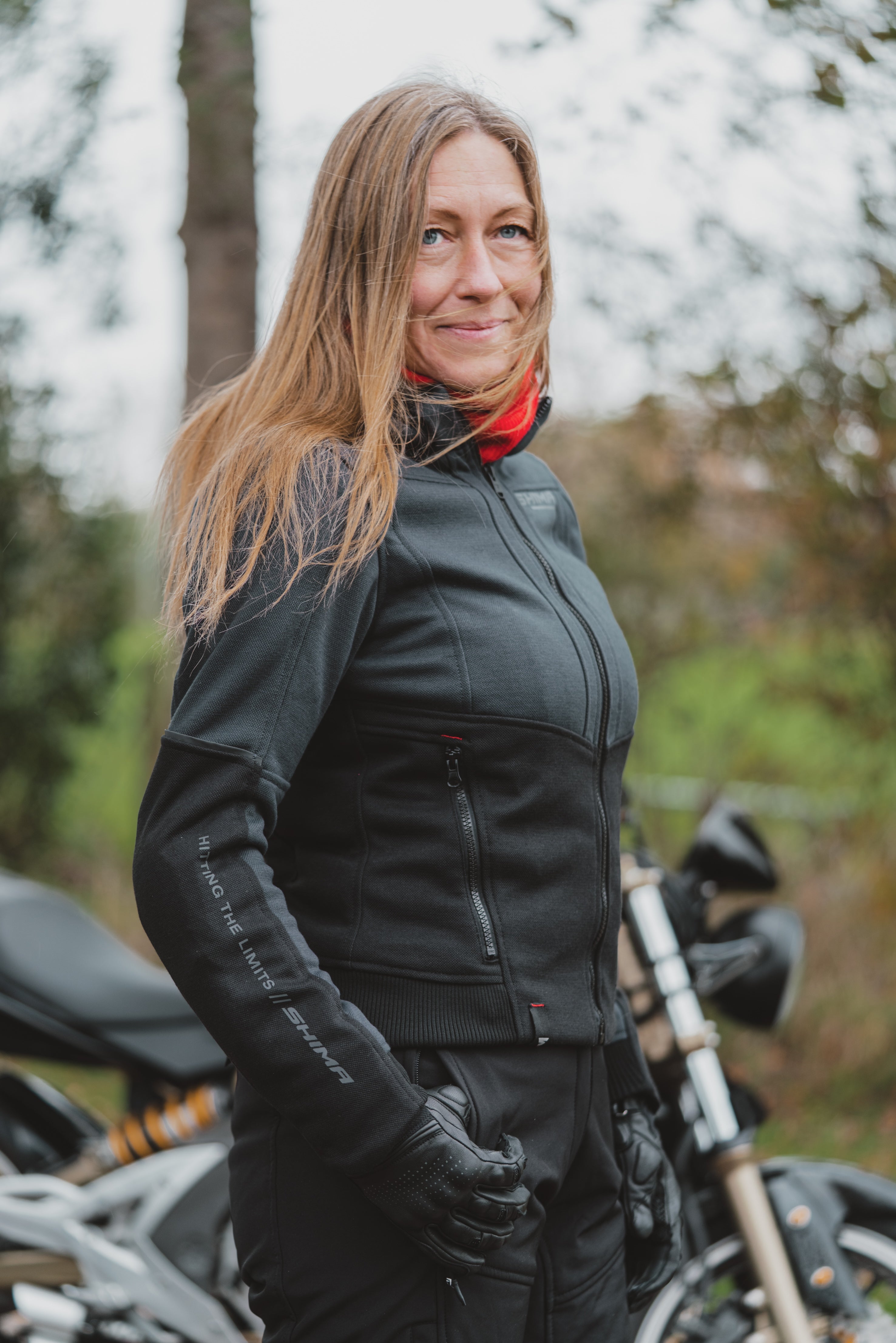 A woman wearing a black motorcycle jacket, matching black pants, and black gloves, standing outdoors next to a motorcycle with a blurred forest background.