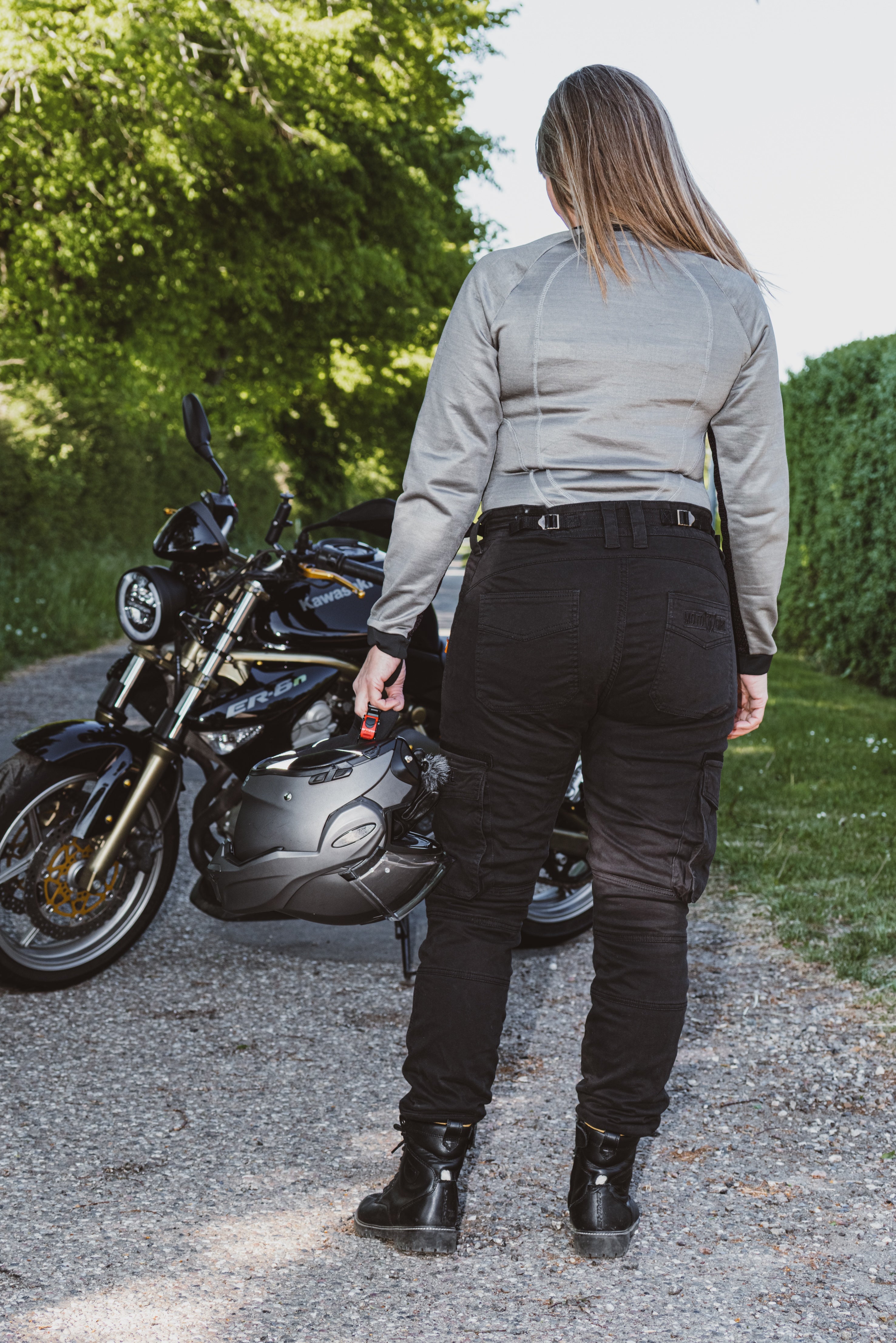 Person standing next to a motorcycle with a helmet, wearing a light gray shirt and black pants.