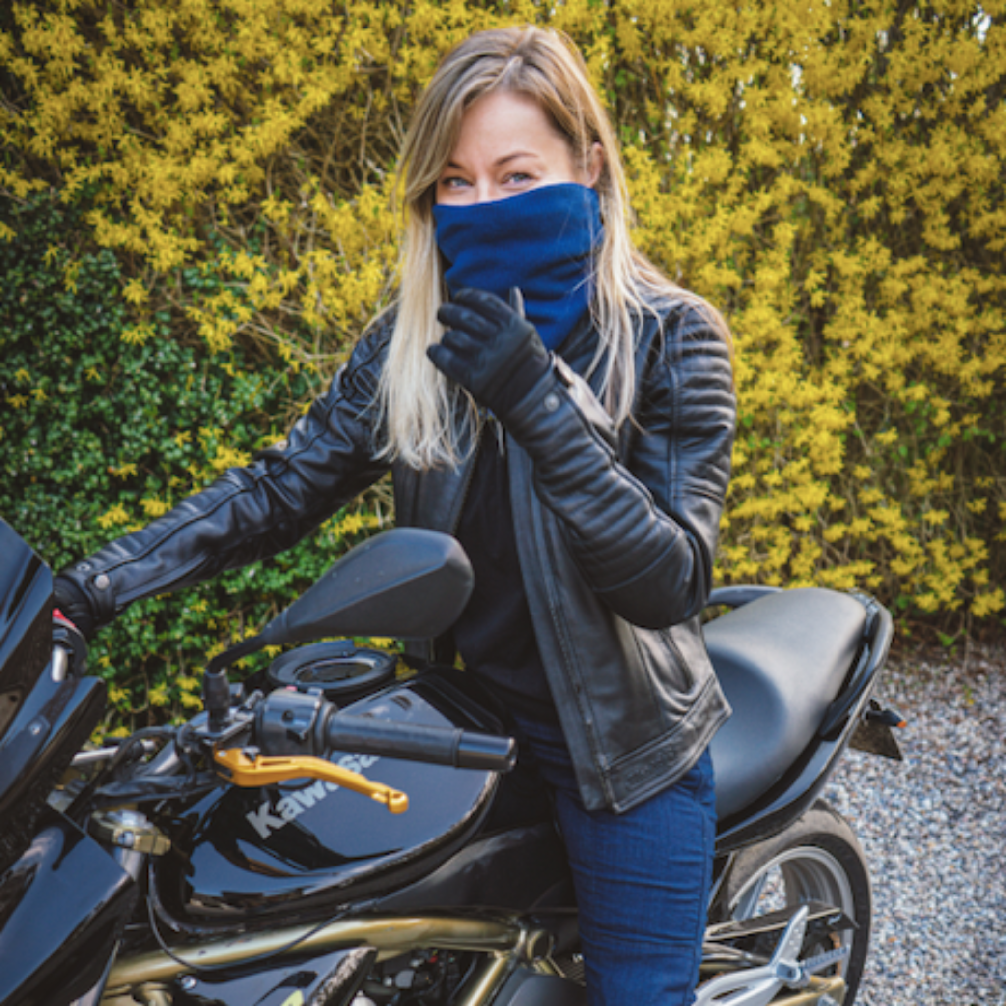 young light haired woman sitting on a Kawasaki motorcycle while wearing blue knitted neck warmer from Moto Girl and black Valerie jacket.