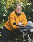 Smiling women sitting on a motorcycle surrounded by nature, wearing a yellow Valerie jacket and a green knitted neck warmer from Moto Girl.