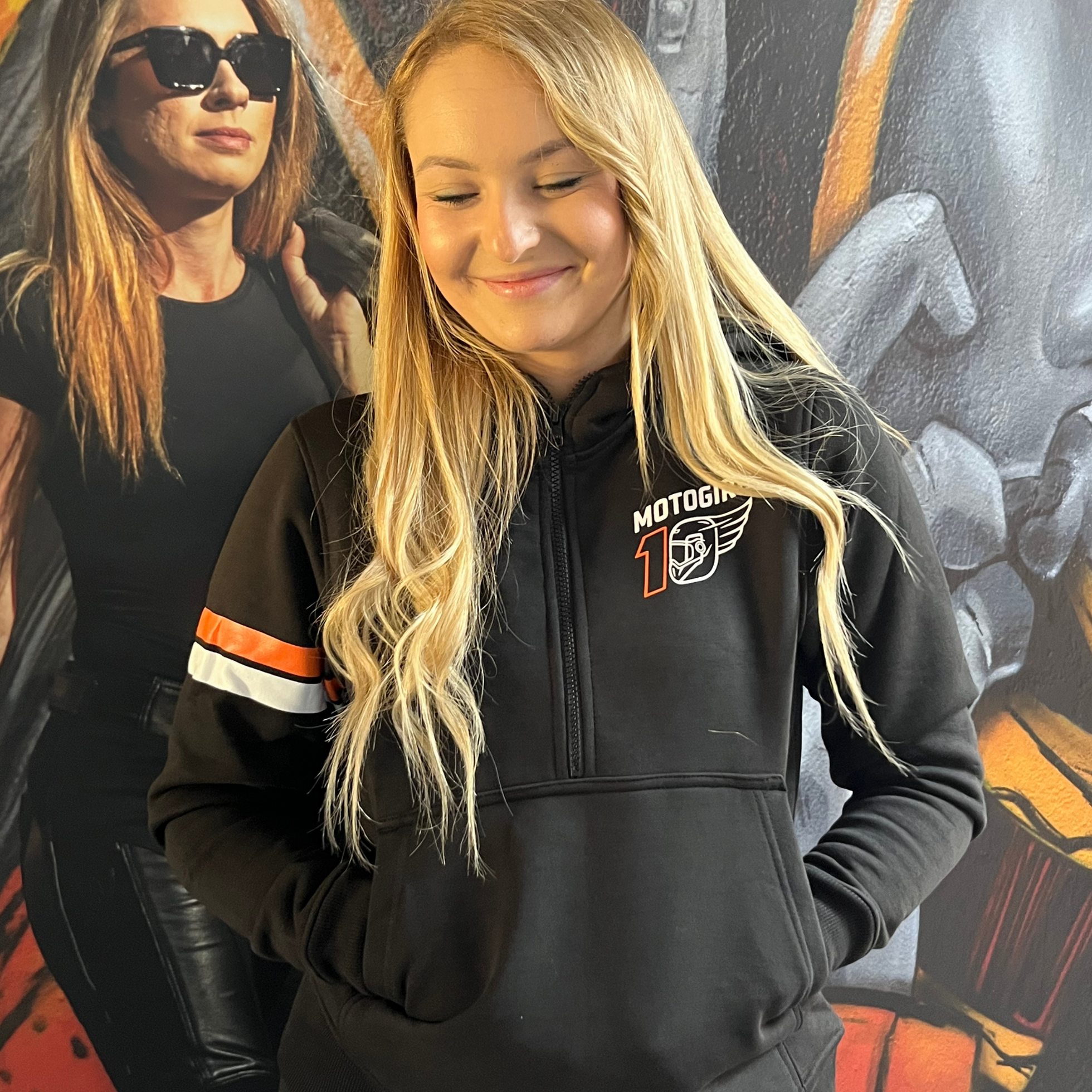 Person wearing a black hoodie with a logo, standing in front of a colorful mural.