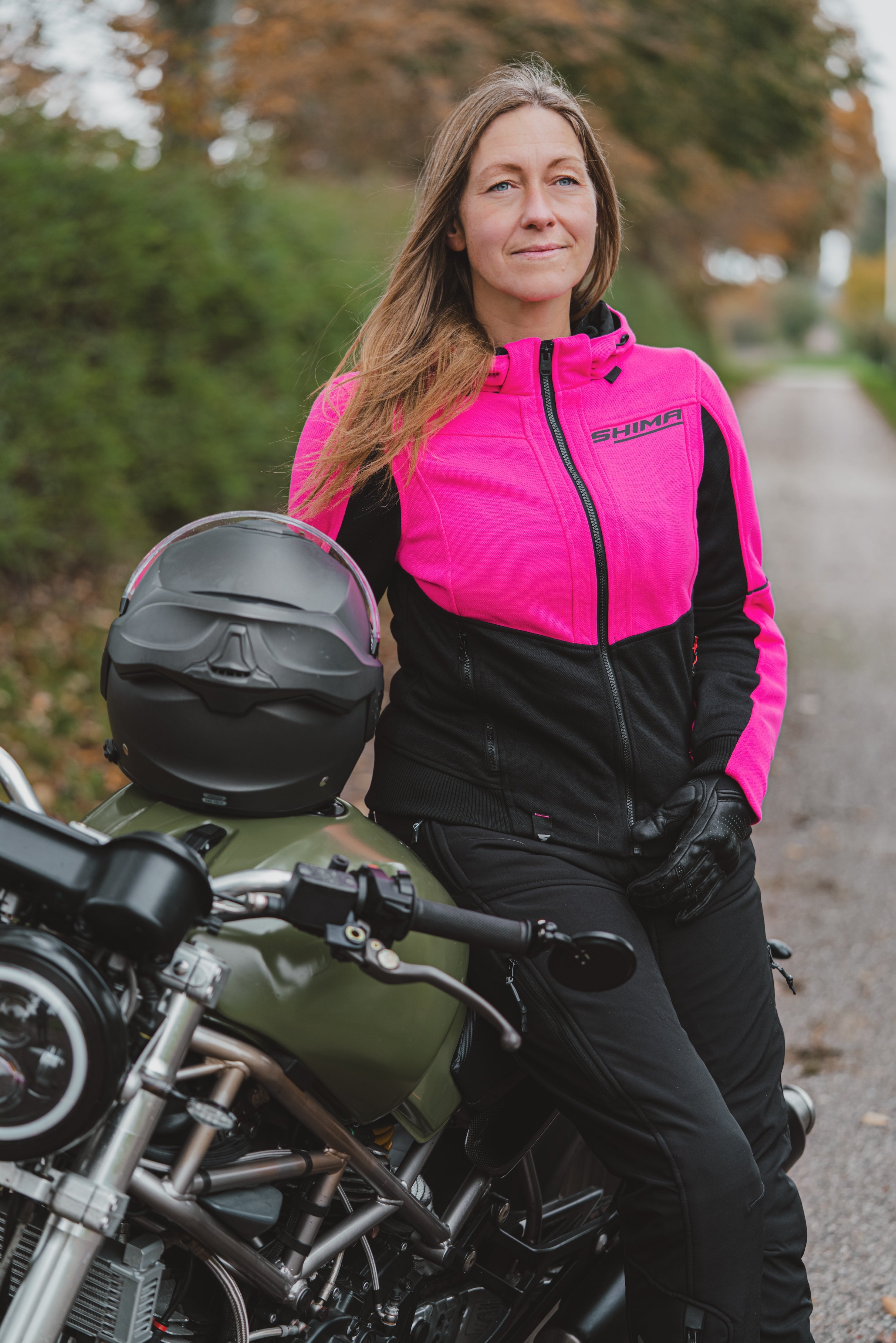 A woman wearing a bright pink and black motorcycle jacket sits on an olive green cruiser motorcycle outdoors. A black helmet rests on the motorcycle seat, and trees and a road are blurred in the background.