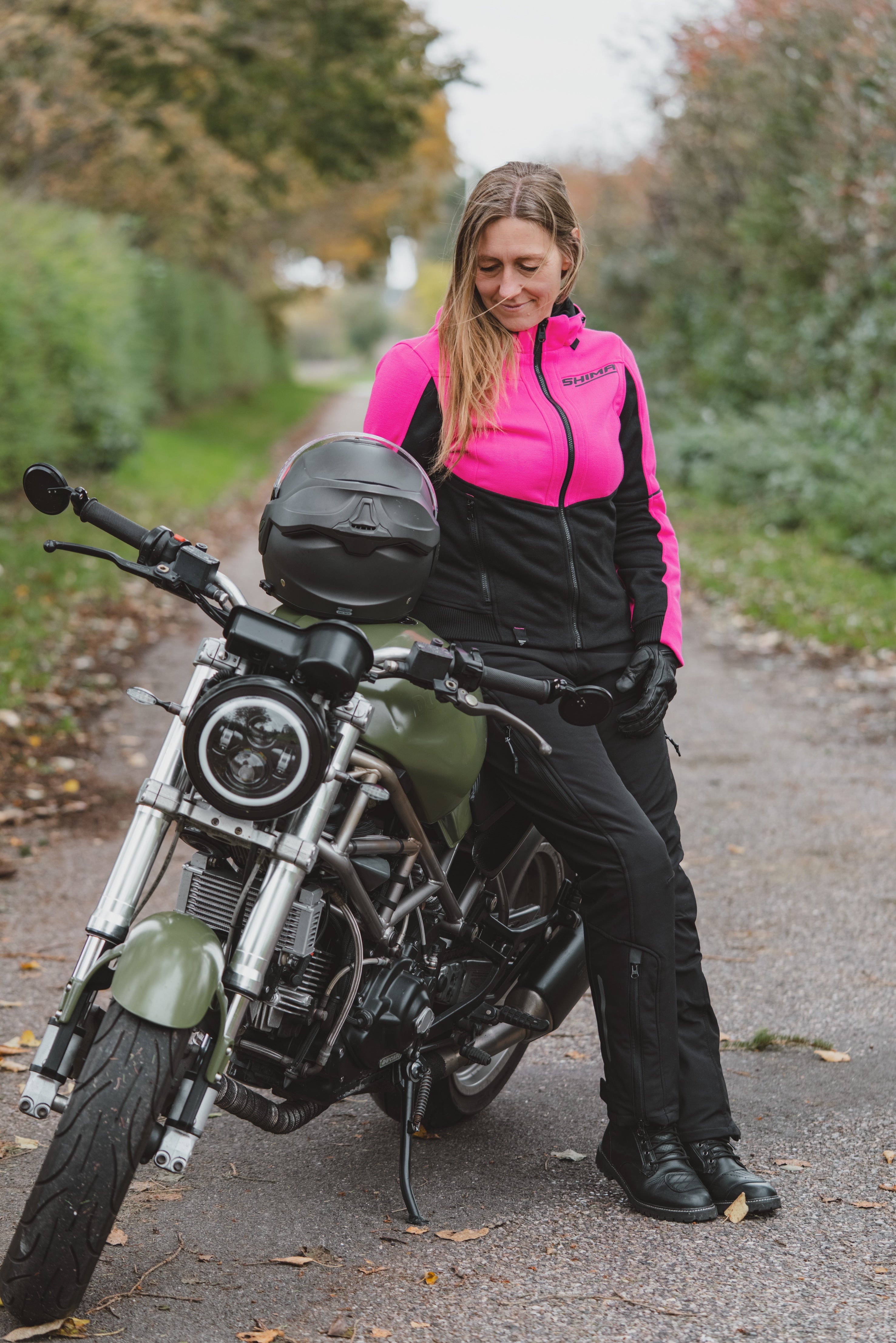 A woman wearing a bright pink and black motorcycle jacket, black pants, and gloves sits on a cruiser-style motorcycle on an outdoor path.