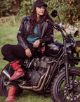 A woman leaning on her Triumph motorcycle wearing black leather motorcycle jacket and red boots 