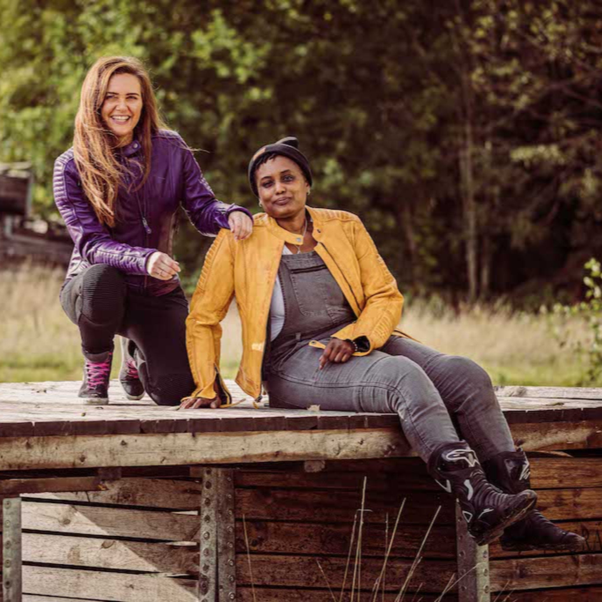 two smiling women sitting outside wearing a mustard yellow and purple Valerie leather jacket from moto girl