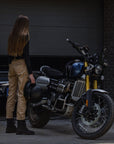 A brunette woman wearing beige Cargo Mila pants, standing next to her motorcycle and holding her helmet, viewed from the back.